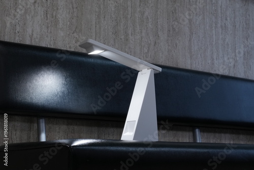 Modern desk lamp rests on sleek black surface. Its minimalist design highlights clean lines and cool tones. Soft light illuminates workspace with subtle, focused glow