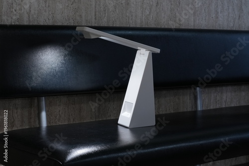Minimalist lamp rests on sleek black bench. Modern design emits soft, focused light. Clean lines suggest contemporary style. Ideal for study, work, or quiet reading