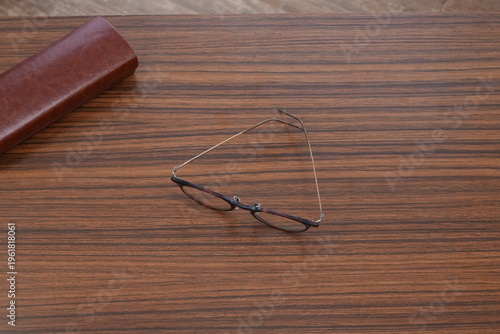 Glasses rest on a warm wooden surface. Brown case lies nearby, unused and still. Metal frames gleam subtly against wood grain. Stillness speaks of quiet contemplation or pause