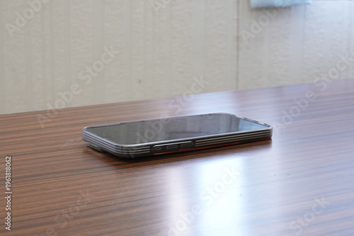 Smartphone rests on polished wooden surface. Glossy screen reflects surrounding light subtly. Soft background features textured wall panels. Minimalist scene suggests quiet, modern simplicity