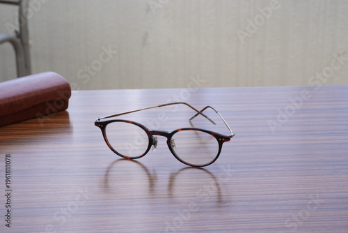 Elegant round glasses rest on polished wooden table. Soft lighting highlights gentle curves and reflective surfaces. Brown leather case nearby adds touch of sophistication