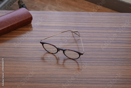 Vintage glasses rest on a warm wooden table. Soft light casts gentle shadows around the frame. Brown leather case lies nearby, suggesting care and use. Minimalist setting evokes quiet nostalgia