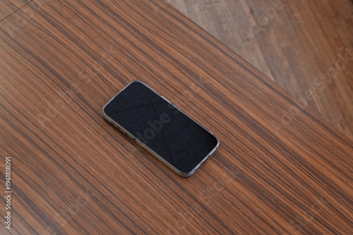 Black phone rests on wooden surface. Simple, modern design with dark screen. Texture contrasts between device and grain. Quiet stillness suggests minimalism or pause. Ideal for tech, lifestyle