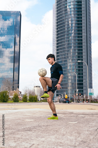 Young man demonstrating soccer skills, balancing and juggling a football in a modern city street with skyscrapers