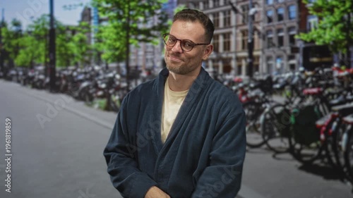 Man wearing robe and glasses pointing with right hand toward bicycles on a crowded street in front of brick buildings, slight smile and folded left arm; amused nonchalance.