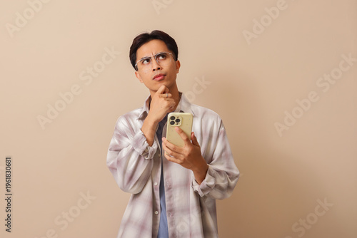 Young Asian man using mobile phone while holding chin and looking up with thoughtful expression isolated on brown background.