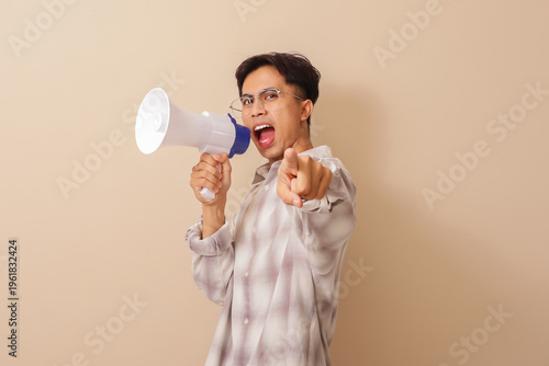 Photos Excited young Asian man shouting into a megaphone with clenched fist, celebrating victory and success in a lively studio portrait, showing enthusiasm and motivation