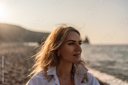 Woman beach sunset, beautiful young blonde woman gazing at the ocean during golden hour on a serene pebble beach.