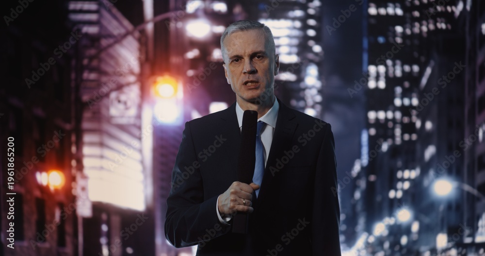 Fototapeta premium Serious Mature Man Journalist Delivering Live Report, Holding Microphone and Looking at Camera. Concept of Media Correspondence, Professional Portrait of Reporter Against Blurred City Night.
