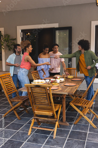 Diverse friends standing and leaning around wooden dining table on tiled patio, passing plates