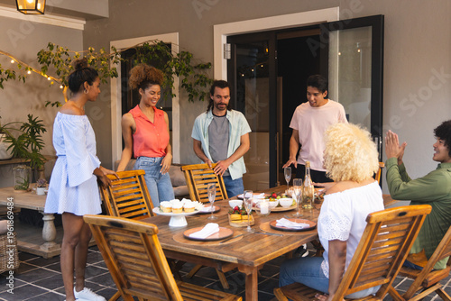 Diverse friends gathering around wooden table on covered patio, sharing cupcakes and sparkling wine