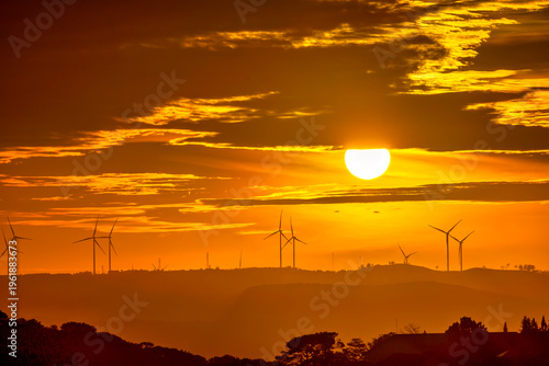 Dramatic sunset scene featuring glowing sun descending behind a horizon lined with wind turbines. The sky is painted in intense golden and orange hues, with layered clouds partially veiling the sun.