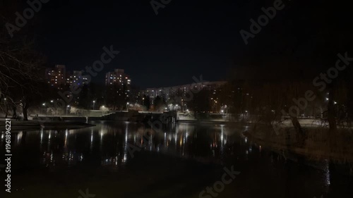 City lights reflecting in calm lake at night, quiet residential waterfront scene with apartment buildings, urban park atmosphere and moody evening skyline background.