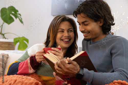 Couple sitting closely on sofa, holding red mug and reading red hardcover book at home