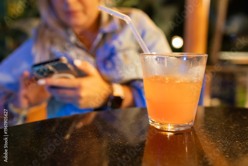 Close-up of an orange cocktail with ice and straw on a table with a blurry woman using smartphone at a night bar