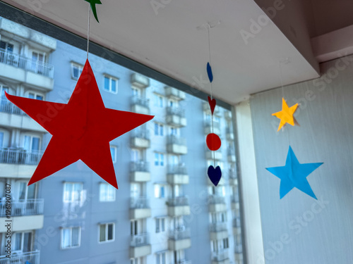 Colorful Paper Stars and Shapes Hanging Ornament Decorations in Front of Window and Apartment Building Background
