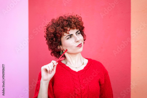 Curly redhead in red sweater thinking with pencil against vibrant background