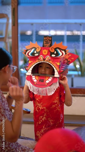 Thai mother and young daughter in traditional red Qipao playing with DIY Chinese dragon mask during Lunar New Year celebration at home.