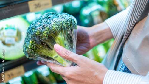 Фотография Close-up of a shopper's hand holding fresh broccoli wrapped in plastic film on a supermarket shelf