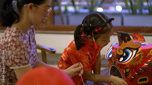 Thai mother and young daughter in traditional red Qipao playing with DIY Chinese dragon mask during Lunar New Year celebration at home.