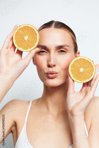 Young woman with clear skin holding two halves of fresh orange fruit near her face. Natural beauty concept with healthy lifestyle and vitamin C. Bright studio lighting, white background.