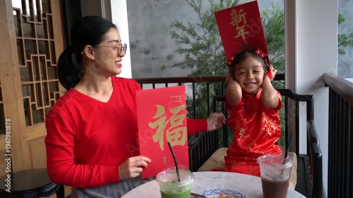 Happy Thai-Chinese mother and daughter in traditional red outfits celebrating Lunar New Year with large red envelopes at an outdoor cafe.