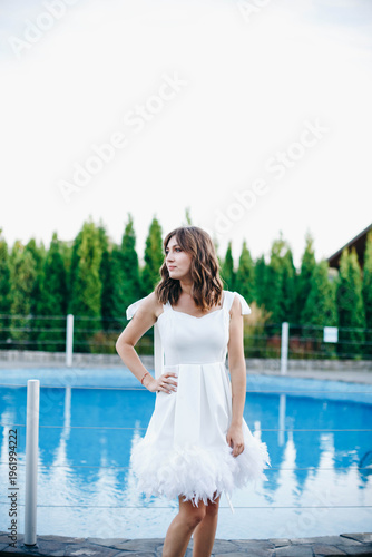 Young woman in white dress with feather hem posing near swimming pool, stylish summer portrait. Elegant feminine look, natural light, modern outdoor lifestyle scene with water reflections.