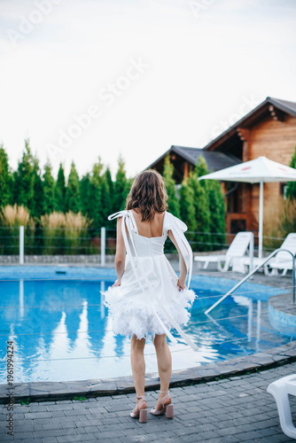 Young woman in white dress with feather hem posing near swimming pool, stylish summer portrait. Elegant feminine look, natural light, modern outdoor lifestyle scene with water reflections.