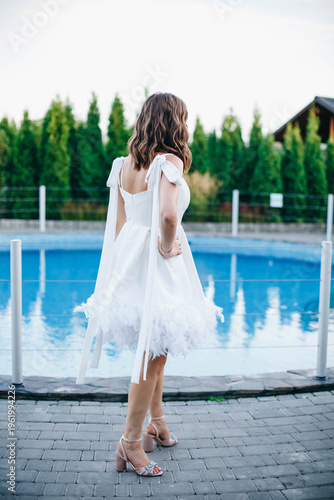 Young woman in white dress with feather hem posing near swimming pool, stylish summer portrait. Elegant feminine look, natural light, modern outdoor lifestyle scene with water reflections.