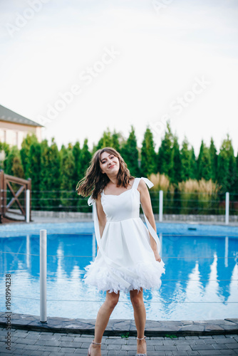 Young woman in white dress with feather hem posing near swimming pool, stylish summer portrait. Elegant feminine look, natural light, modern outdoor lifestyle scene with water reflections.
