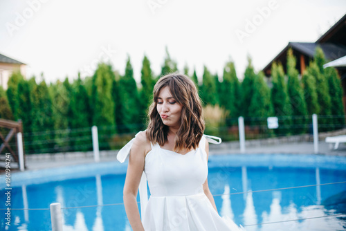 Young woman in white dress with feather hem posing near swimming pool, stylish summer portrait. Elegant feminine look, natural light, modern outdoor lifestyle scene with water reflections.