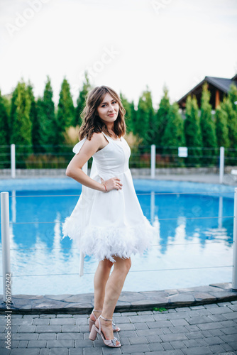 Young woman in white dress with feather hem posing near swimming pool, stylish summer portrait. Elegant feminine look, natural light, modern outdoor lifestyle scene with water reflections.