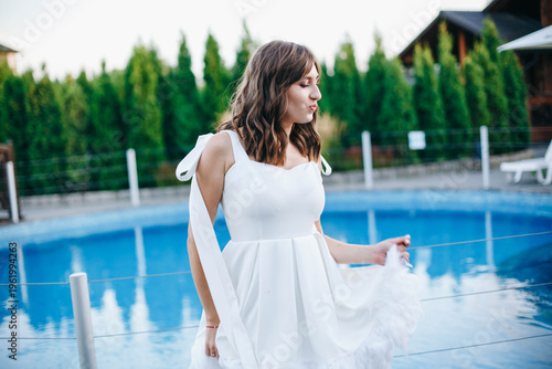 Young woman in white dress with feather hem posing near swimming pool, stylish summer portrait. Elegant feminine look, natural light, modern outdoor lifestyle scene with water reflections.