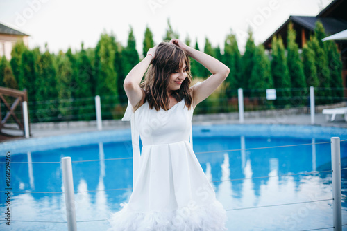 Young woman in white dress with feather hem posing near swimming pool, stylish summer portrait. Elegant feminine look, natural light, modern outdoor lifestyle scene with water reflections.