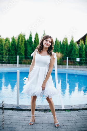 Young woman in white dress with feather hem posing near swimming pool, stylish summer portrait. Elegant feminine look, natural light, modern outdoor lifestyle scene with water reflections.