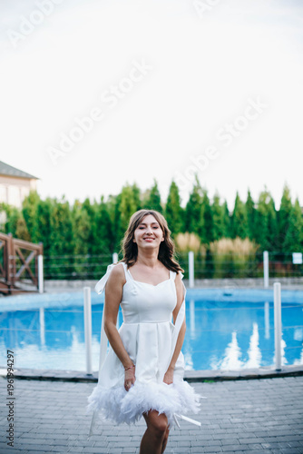 Young woman in white dress with feather hem posing near swimming pool, stylish summer portrait. Elegant feminine look, natural light, modern outdoor lifestyle scene with water reflections.