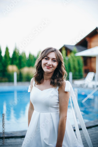 Young woman in white dress with feather hem posing near swimming pool, stylish summer portrait. Elegant feminine look, natural light, modern outdoor lifestyle scene with water reflections.
