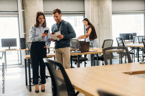 Wallpaper Mural Two professionals discussing work while using mobile devices in a modern office space with desks and computers arranged in the background Torontodigital.ca