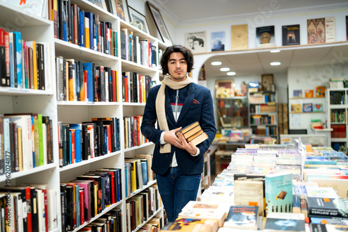 Принти на полотні Young man holding books in a second-hand bookstore aisle