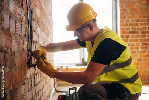 Male electrician wearing yellow safety vest and hard hat works on electrical wiring against a brick wall in a construction site with natural light coming through a window