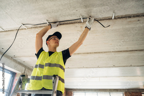 Male electrician in safety vest installing electrical wiring on ceiling while standing on a ladder inside a modern building under construction with large windows
