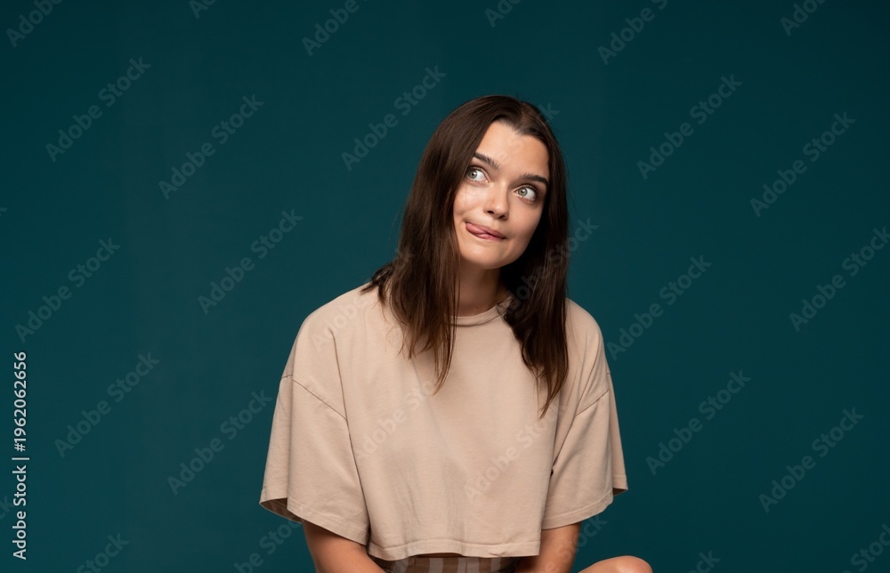 Obraz premium Young woman making a goofy facial expression with tongue out, looking sideways, playful and humorous on teal background