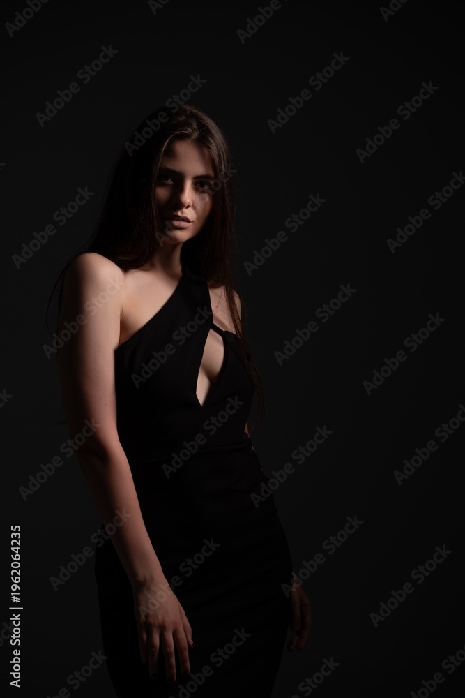 Naklejka premium Confident woman in a one-shoulder black evening dress with keyhole cutout, posing against a dark background; low-key lighting adds mystery and elegant, sophisticated glamour