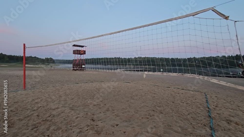 Volleyball net is set up on a sandy beach. The sky is blue and the sun is setting, moving shot
