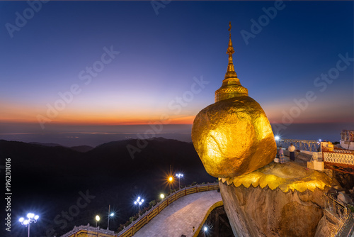 Golden Rock Pagoda Kyaiktiyo Myanmar at twilight. Illuminated balancing boulder pagoda shines brightly against sunset sky & dark hills. View with path, lights, and pilgrims. Iconic sacred site.