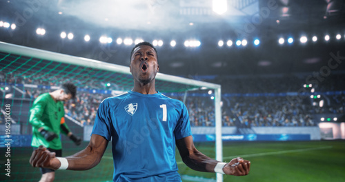 Fényképezés Black Male Soccer Player Clenches Both Fists and Shouts in Triumph Beside the Goal After Scoring in an International Championship Match, as the Goalkeeper Lowers His Head in a Packed World Stadium