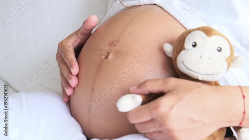 Expectant mother holding a stuffed monkey toy gently stroking her pregnant belly in bed