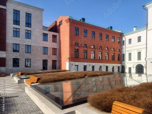 Modern urban courtyard with wooden benches, glass skylights and colorful building facades under a clear sky.
