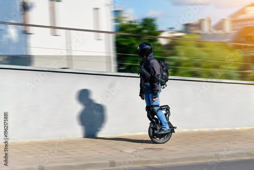 Man riding electric self balancing unicycle in urban environment wearing full protective gear and helmet during sunny day city commute. Micromobility scene, rider on electric unicycle EUC. Motion blur