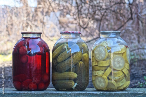 A row of three large glass jars with green pickled cucumbers, zucchini, and red compote with berries stand on a table on a daytime street